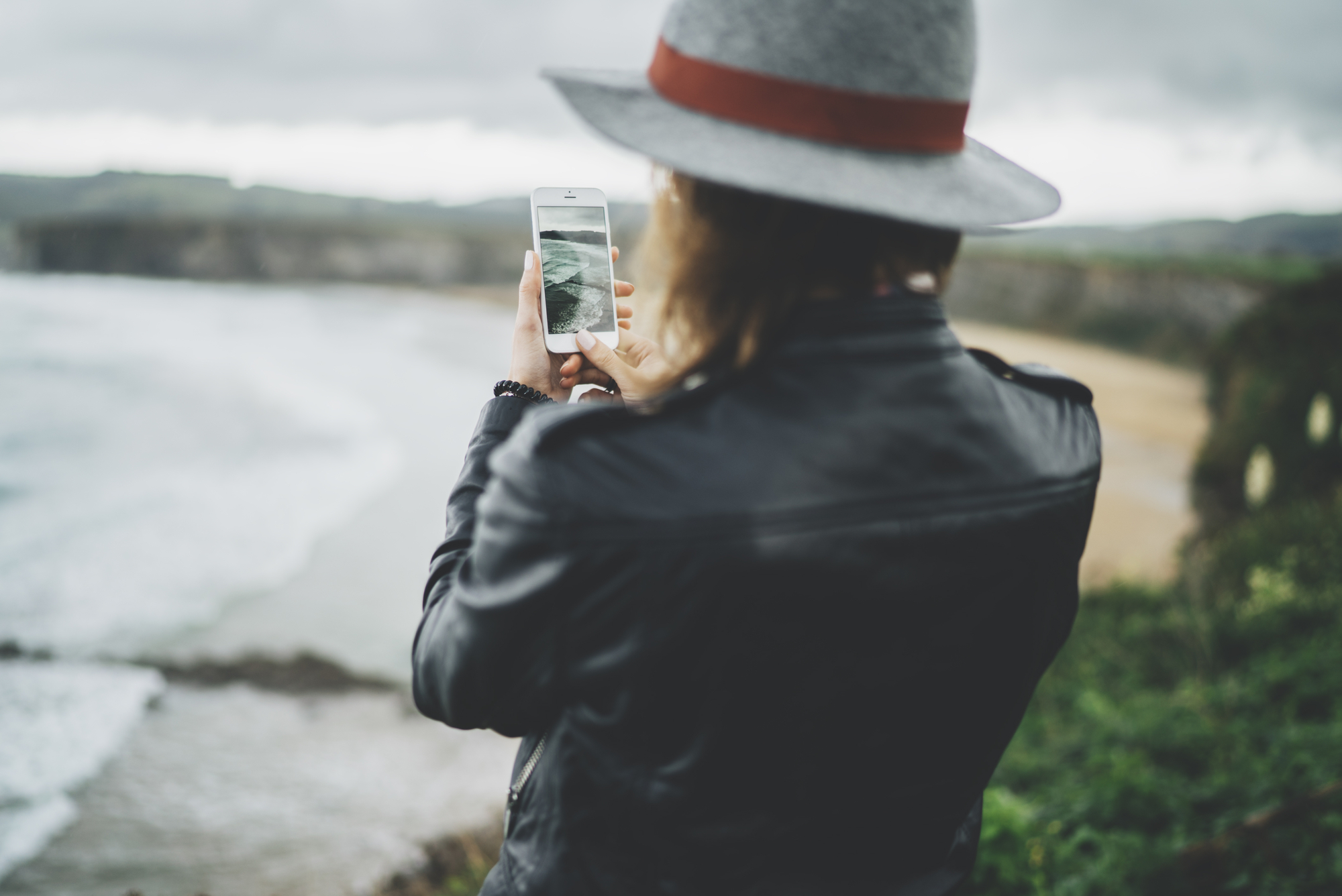 Back view of young traveler girl taking a photo of beautiful ocean via her smartphone device, woman in hipster style is making a photo with cellphone camera an amazing ocean while traveling in Europe, travel and tourism concept